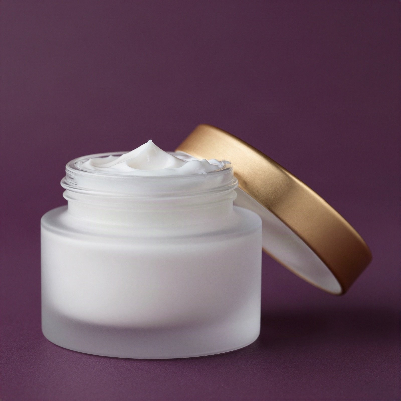 An open frosted glass jar of rich white face cream with a golden lid