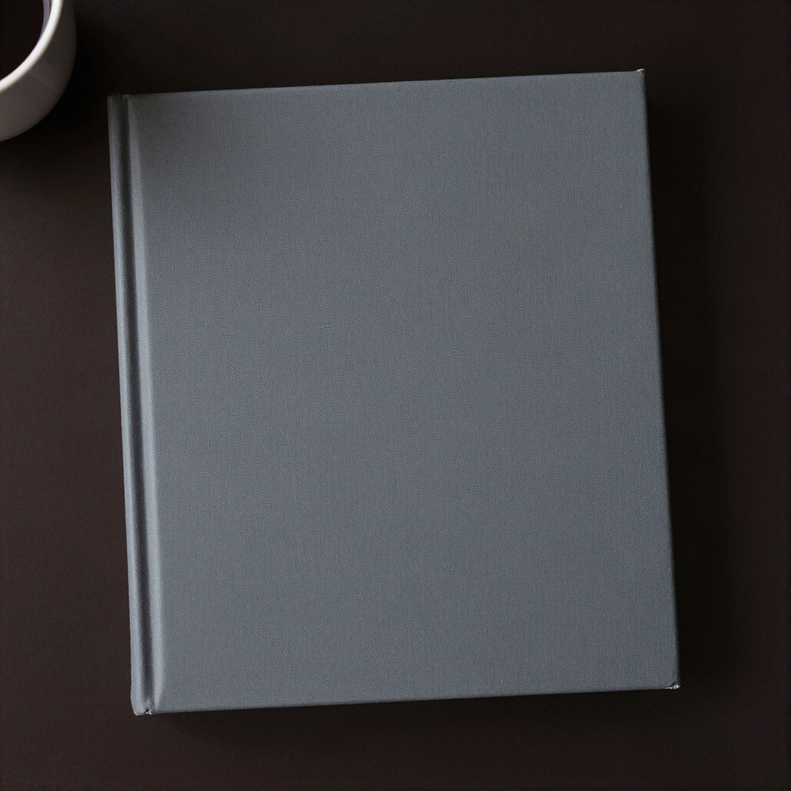 A plain charcoal grey hardcover book on a dark cafe table beside a coffee cup