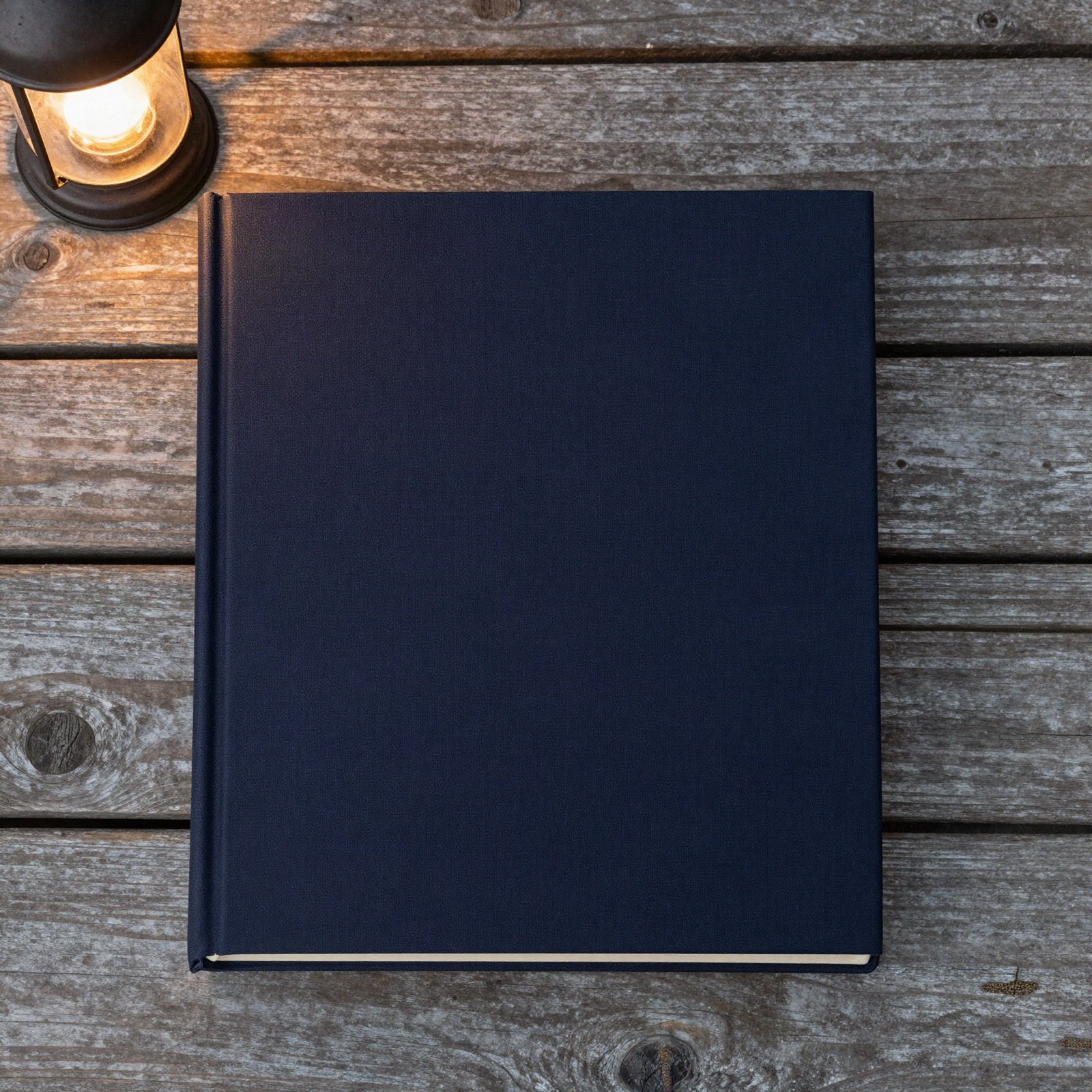 A plain navy hardcover book on weathered porch boards beside a lit lantern
