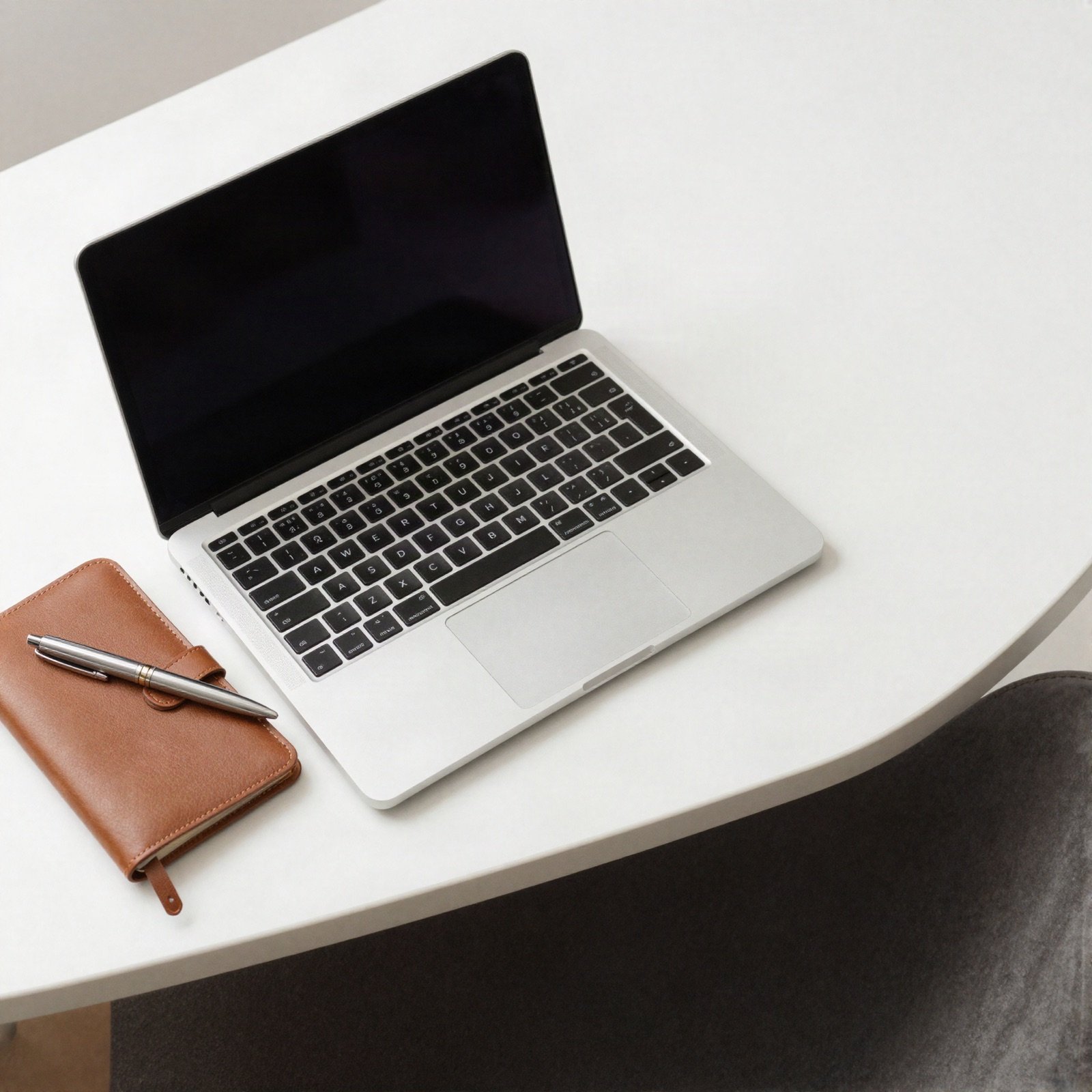 An open laptop beside a leather diary and pen on a white desk