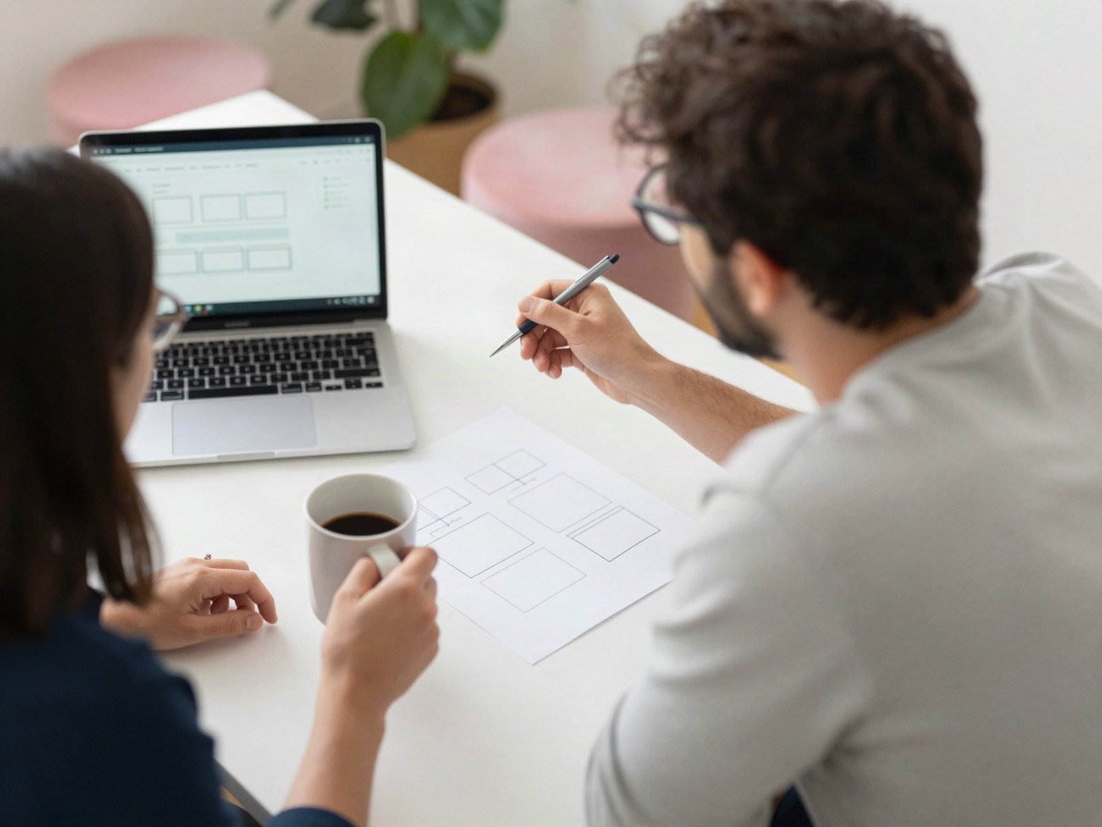 Two web designers planning a page layout with paper sketches and a laptop over coffee