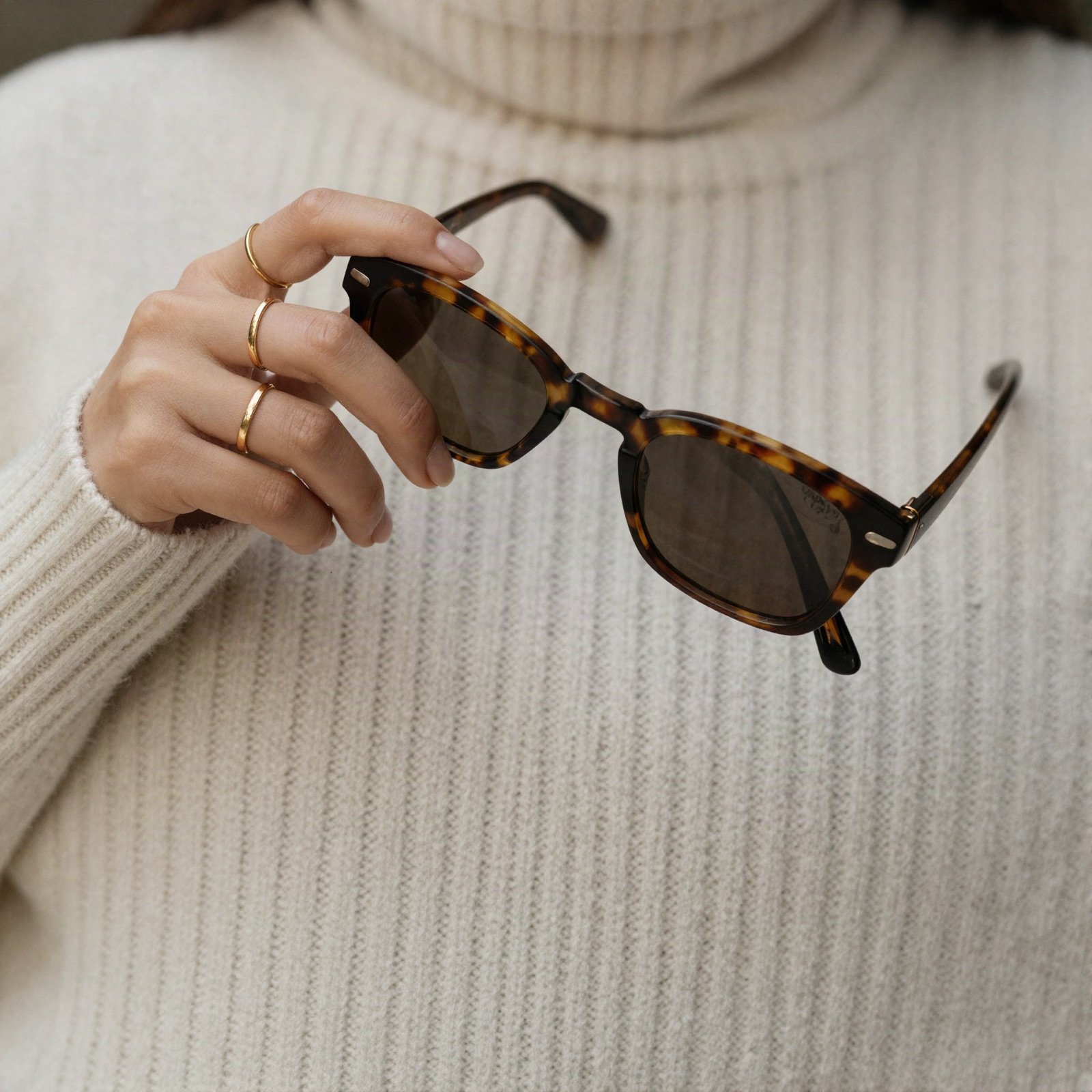 A hand with gold rings holding tortoiseshell sunglasses over a cream sweater