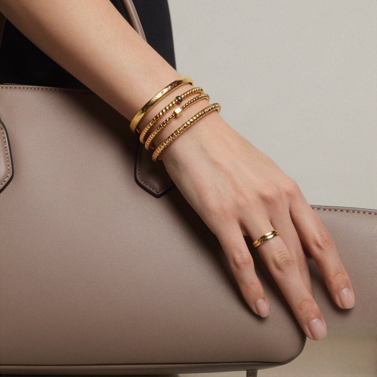 A wrist with layered gold bracelets resting on a taupe leather handbag