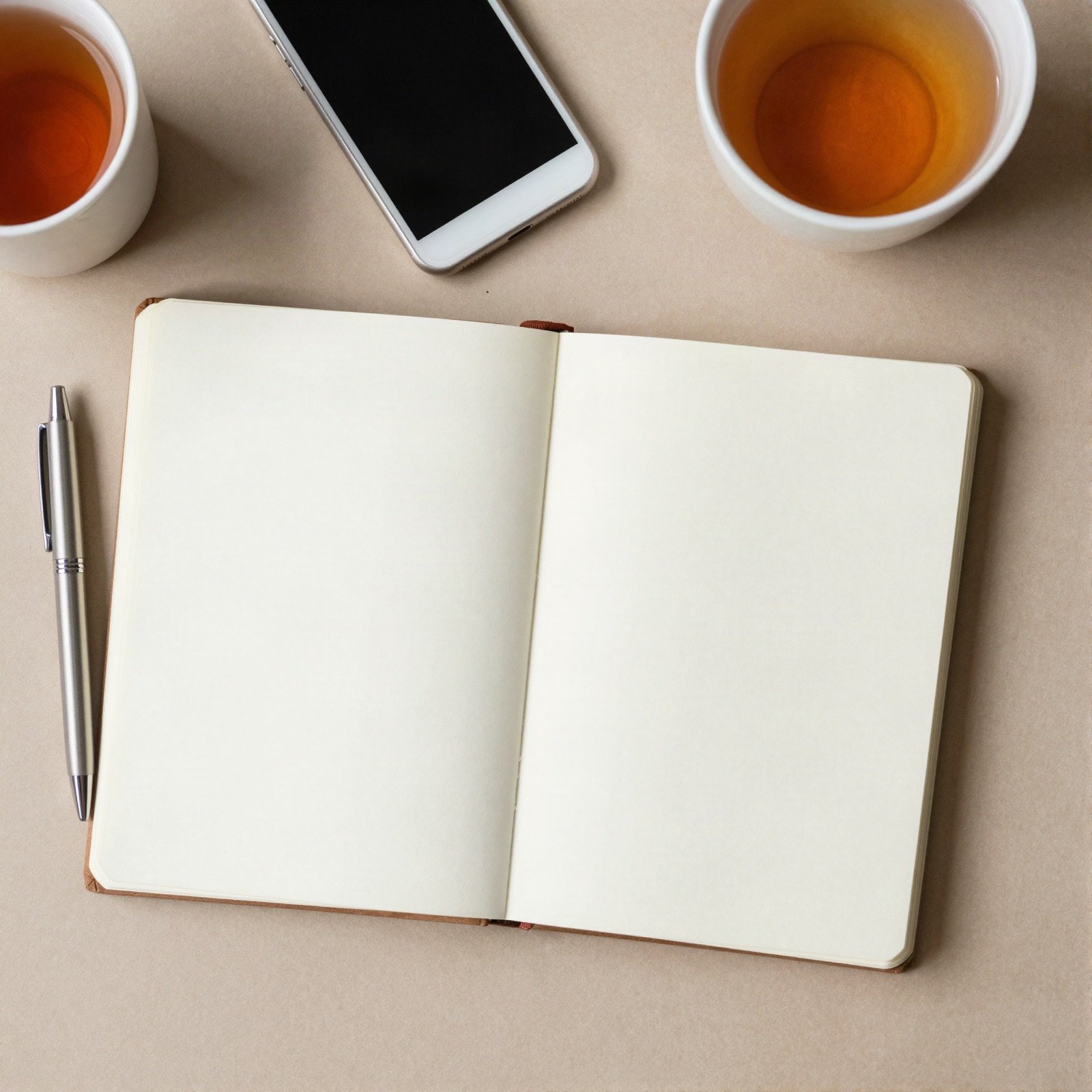 An open blank notebook, a pen, a phone and cups of tea on a desk