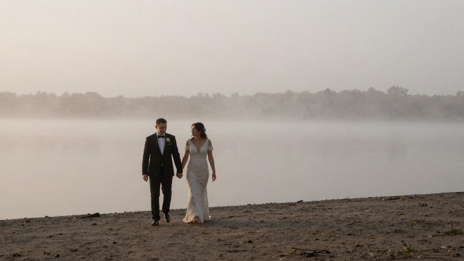 A bride and groom walking along a misty lake shore at dawn